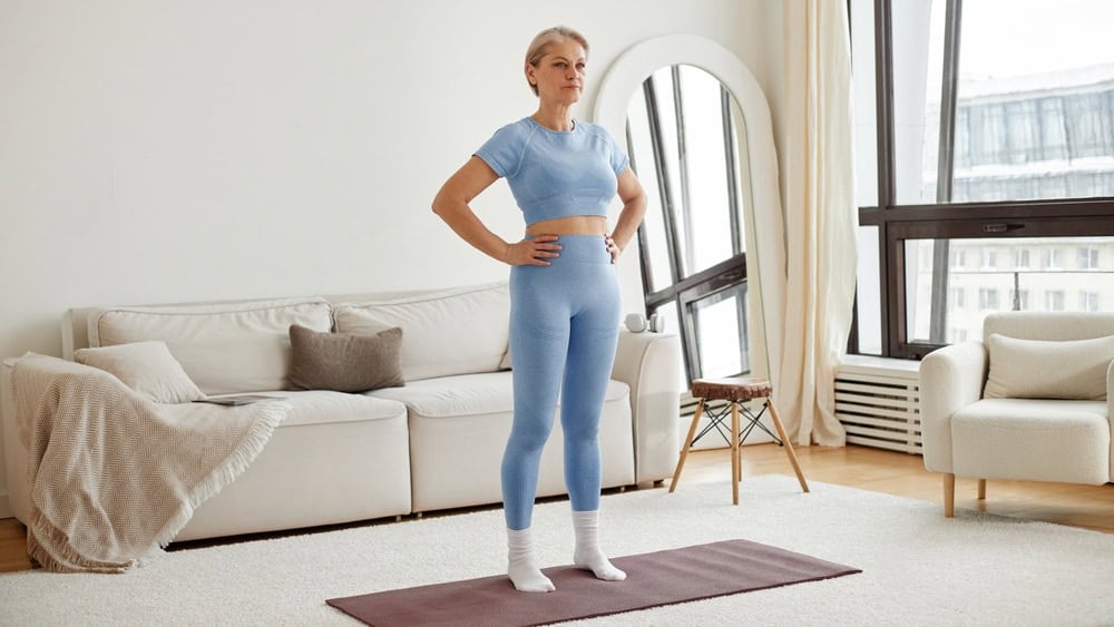 Caucasian mature woman standing on yoga mat with hands on hips preparing for workout in bright living room, looking forward with focused expression, fitness training at home