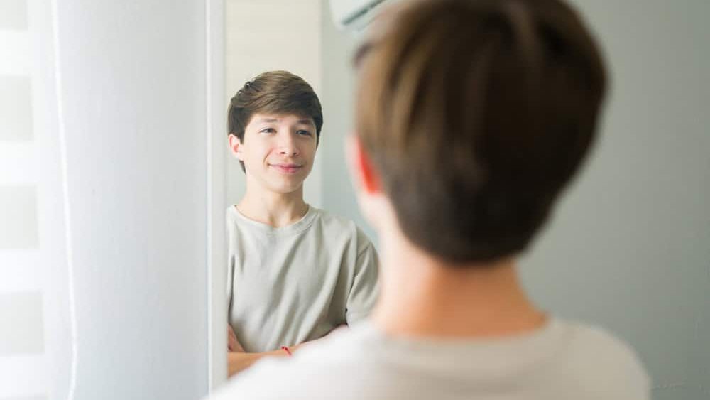 Happy teen boy admiring himself in the mirror with arms crossed, smiling at his reflection in his bedroom