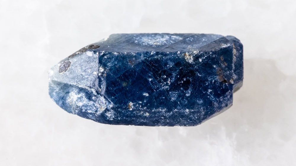 sample of rough single blue sapphire crystal on white marble background close up from Tanzania