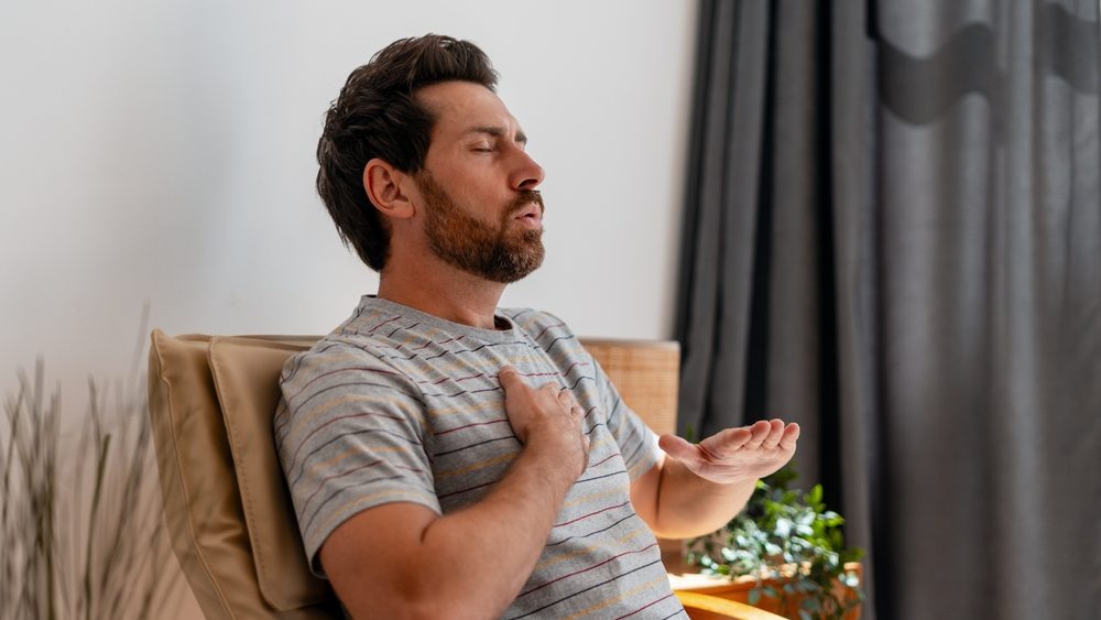 Sad middle aged Latin man experiencing chest pain and breathing difficulty sitting at home in living room. Health care concept