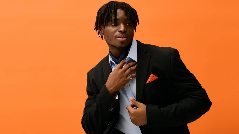 A young, stylish man poses confidently in a black suit jacket with an orange pocket square.