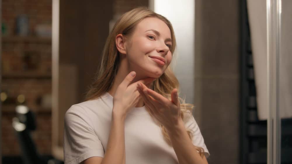 Happy young woman girl applying cream on face skincare look mirror at bathroom Caucasian female model customer touching soft skin antiaging lotion serum massaging neck hands beauty routine cosmetics