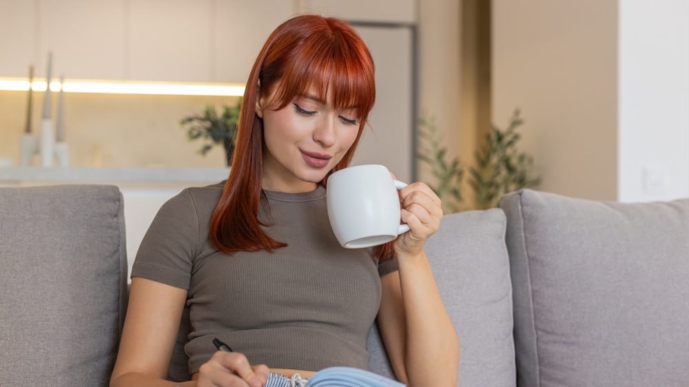 Young woman at home drinks hot tea slowly calms down after long stress and writes task list. Redhead focused girl on sofa holds warm cup writes plan for tomorrow in notebook calm breath and peace
