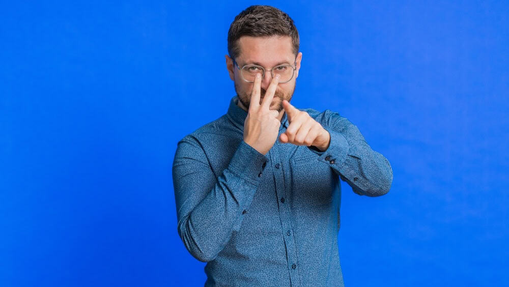 I am watching you. Confident attentive Caucasian young man in glasses pointing at eyes and camera, show I am watching you gesture, spying on someone. Strict adult guy isolated on blue background