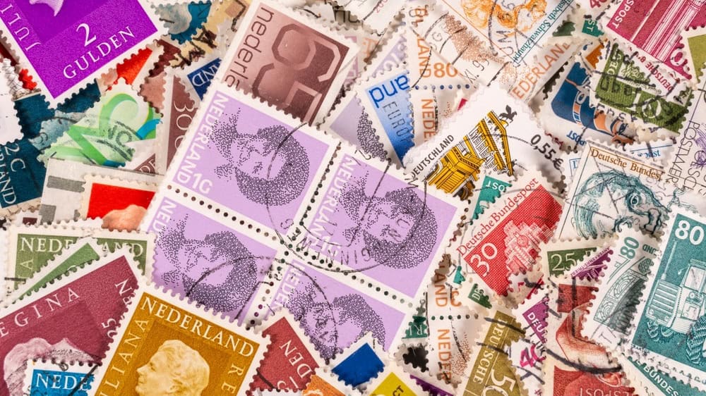 Many stamps scattered on a surface. The stamps are from various countries and years, featuring different designs and colors. Some are used, while others are new.