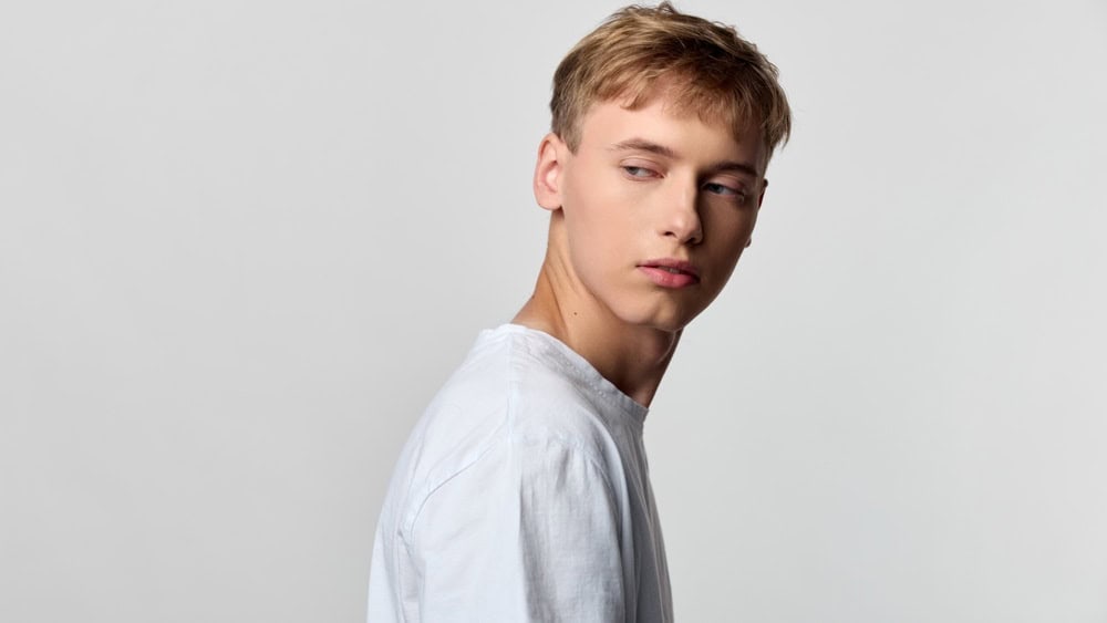 Young man in a plain white shirt turns his head to the side, a calm contemplative expression against a minimal studio backdrop, suitable for fashion, lifestyle, beauty or editorial concepts