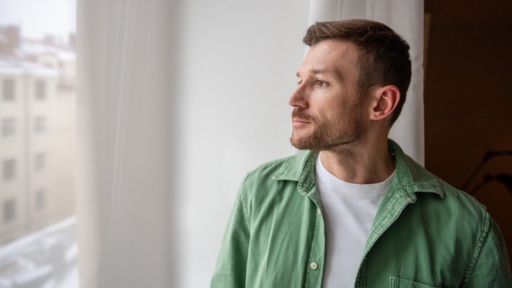 Thoughtful bearded handsome man with focused tranquil expression calmly looking at window, engaged in thinking process, brainstorming new work ideas, reflecting on life and finding inspiration.