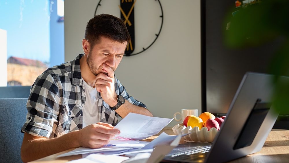 Worried man checking bills and financial papers while sitting at table with laptop. Stressed person planning home budget. Concept of personal finance review, expense control and household budgeting