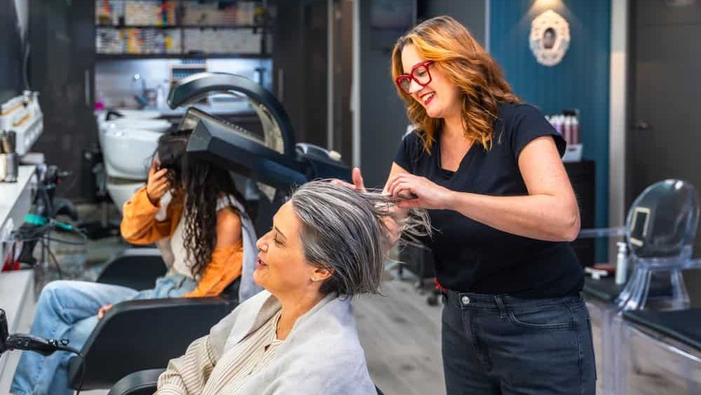 Hairstylist smiling while prepping a senior woman's hair for treatment as another client dries under a hooded dryer in a bright, modern salon setting, relaxed and cheerful