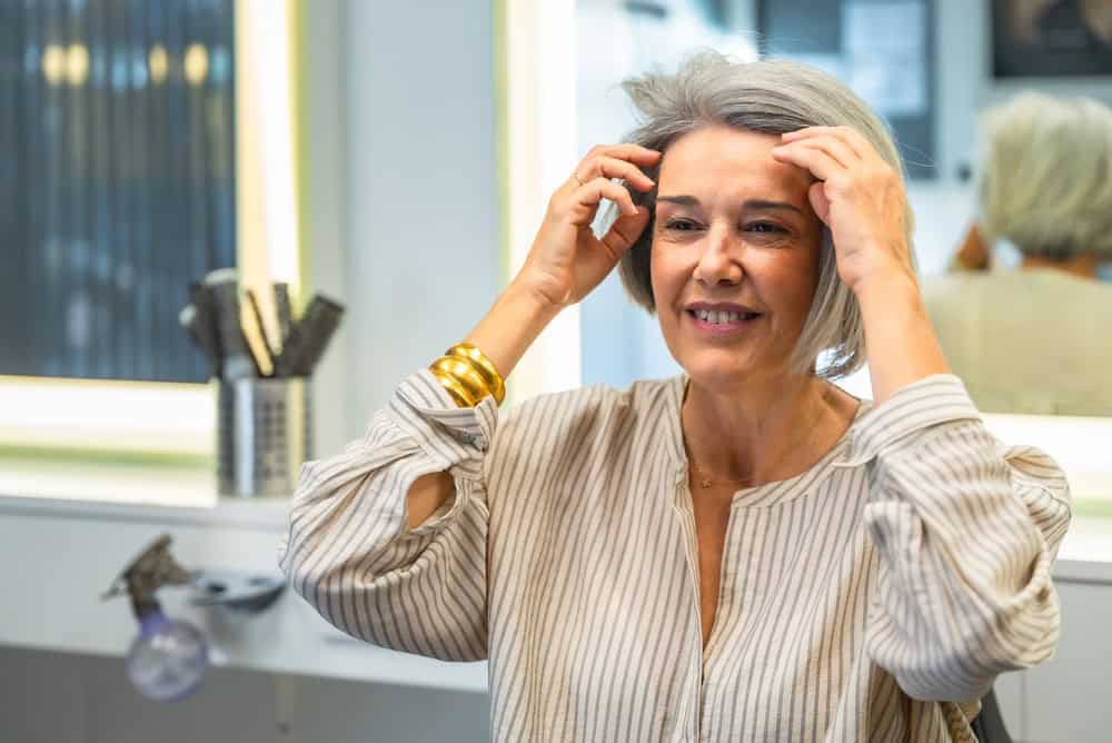 Mature woman looking at her reflection in the mirror, touching her styled gray hair and smiling, feeling happy with her fresh haircut in a modern hair salon environment