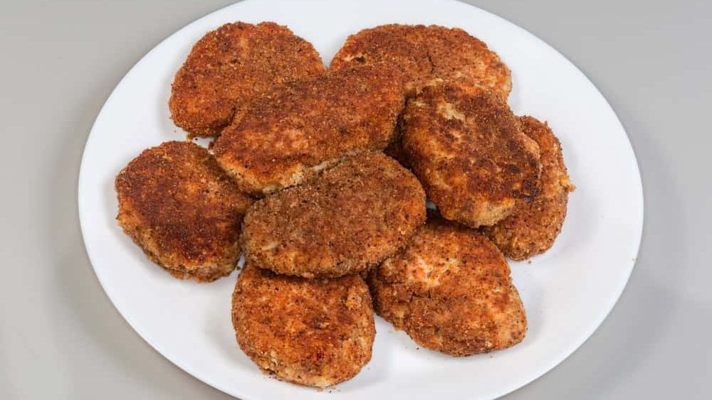 Heap of flat round salmon patties fried in the breadcrumbs on a white plate on a gray background