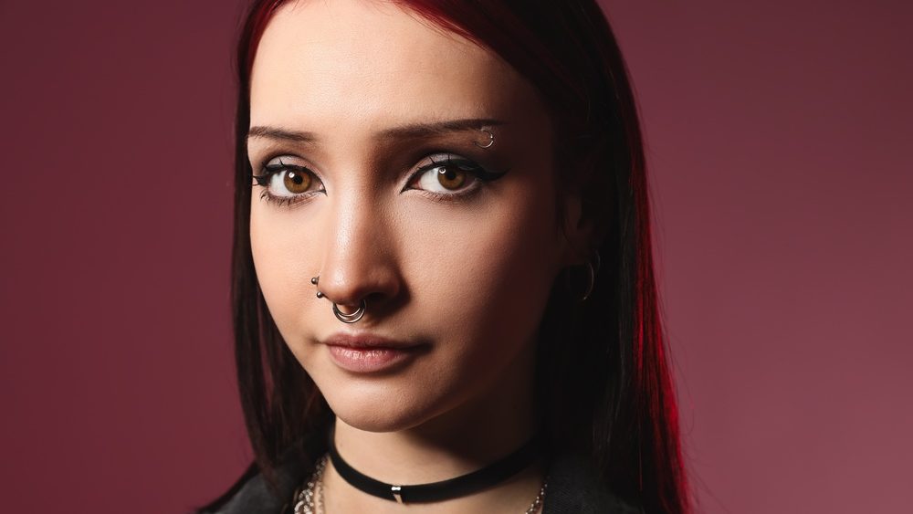 Portrait of beautiful young woman with different face piercings and choker on pink background