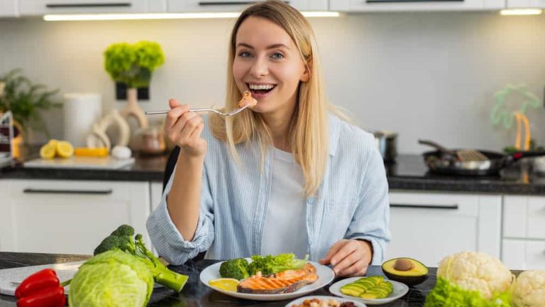 Middle-aged woman eats keto meal with fish trout avocado nuts and greens in home kitchen for healthy weight loss. Girl follows low carb nutrition plan and learns mindful eating for fitness indoors.