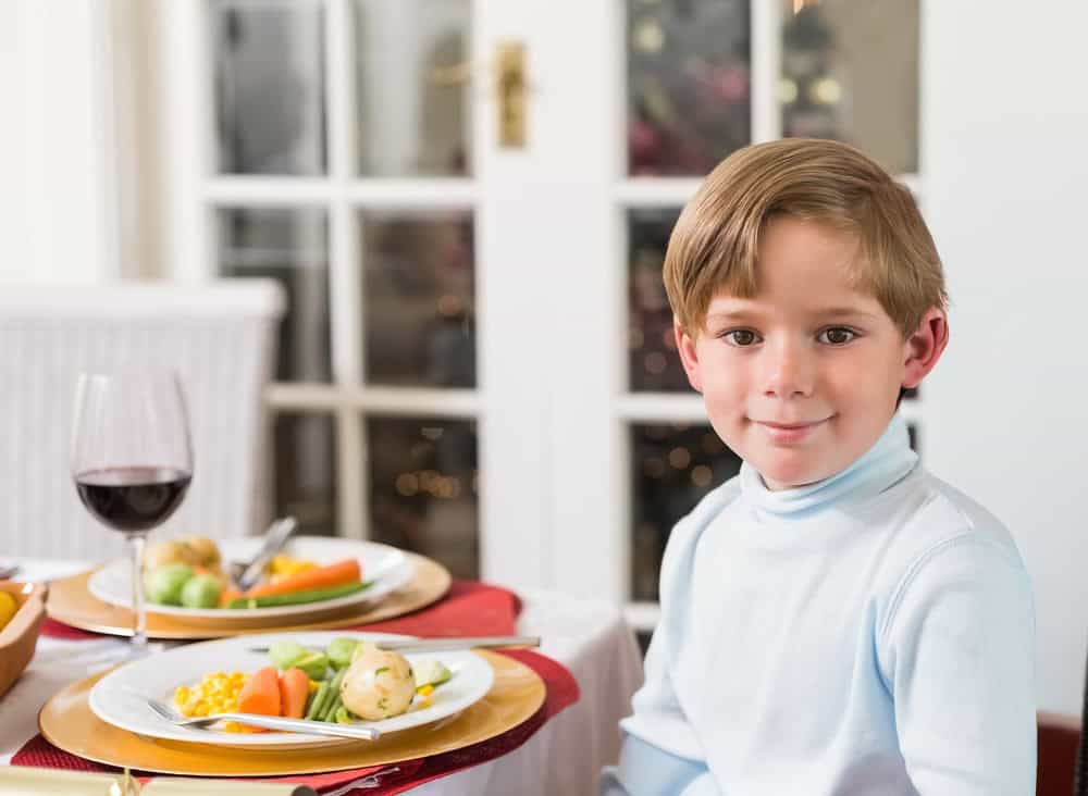 Male child sitting at home dining table wearing light blue turtleneck, vegetables and wine glass. Plates, charger, runner, wicker, brass, doors, warmth