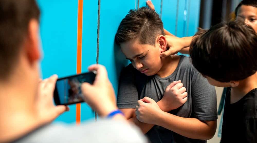 A boy is being bullied by two peers in a school hallway. One is filming the incident on a smartphone. The boy looks distressed and is standing against lockers