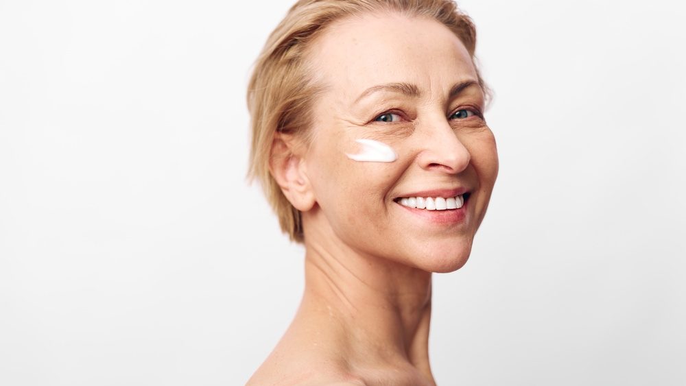 Smiling mature woman with clear skin applies white facial cream on cheek in bright studio. Concept for skincare, anti aging, natural beauty, wellness, and self care with positive mood.