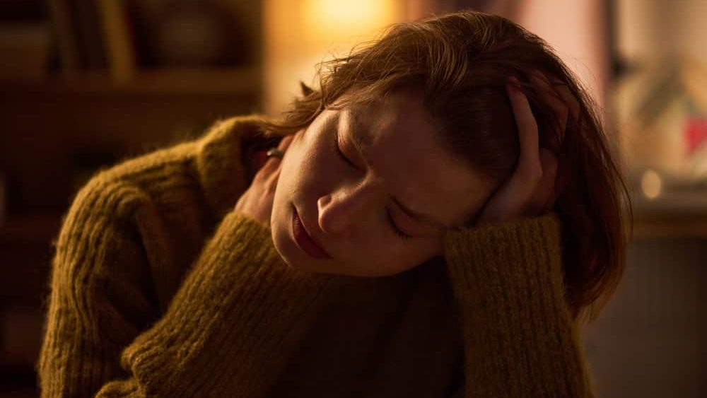 Young woman sitting with head resting on hand, eyes closed and brow furrowed, showing signs of fatigue or discomfort in softly lit indoor setting