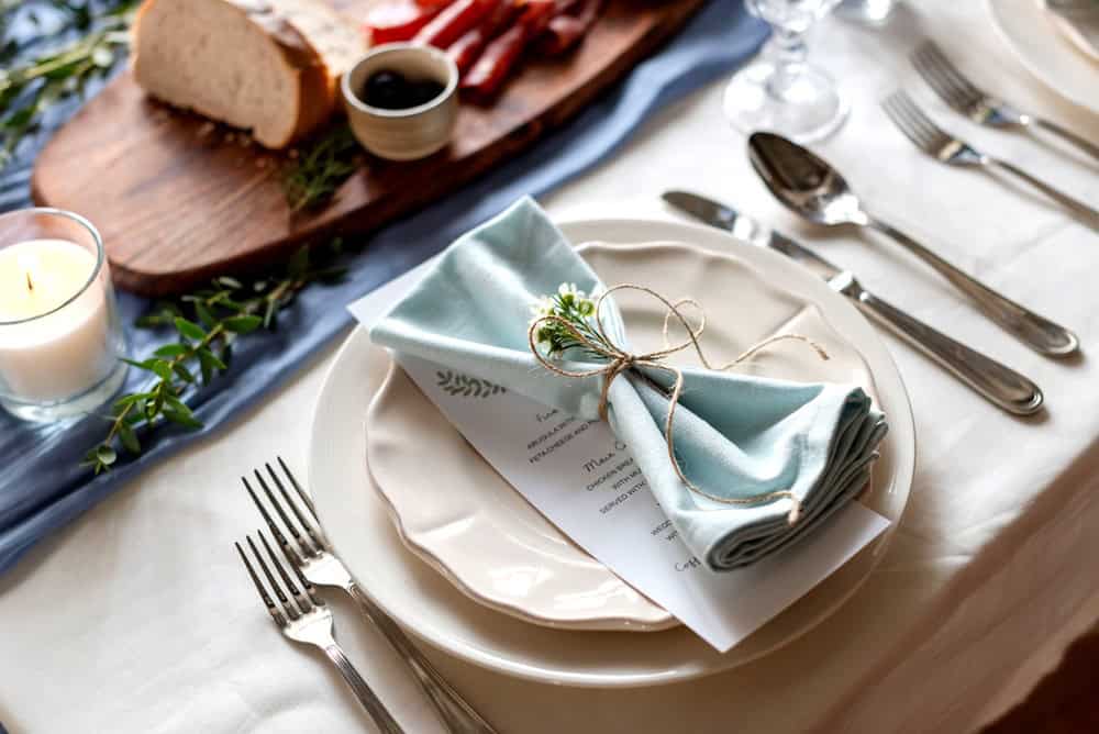 Elegant table setting with a blue napkin, menu card, and greenery on a white plate. Bread and olives on a wooden board in the background.