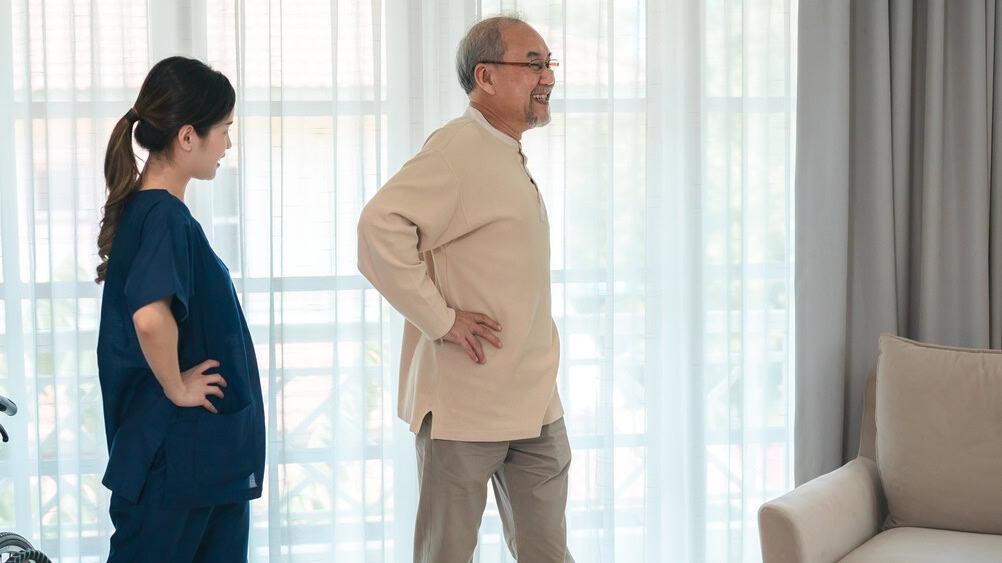 Senior man with caregiver practicing walking exercise in nursing home, focusing on health, insurance, retirement, and mental health support for seniors in comfortable environment