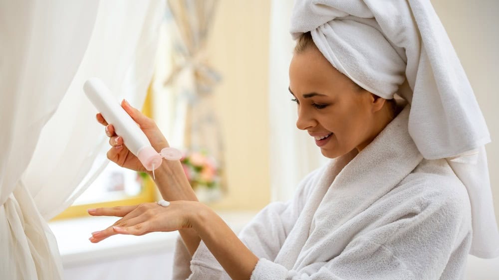 Young woman in bathrobe and towel on head applying skincare lotion on hand in bright bathroom, concept of selfcare, hygiene and beauty routine.