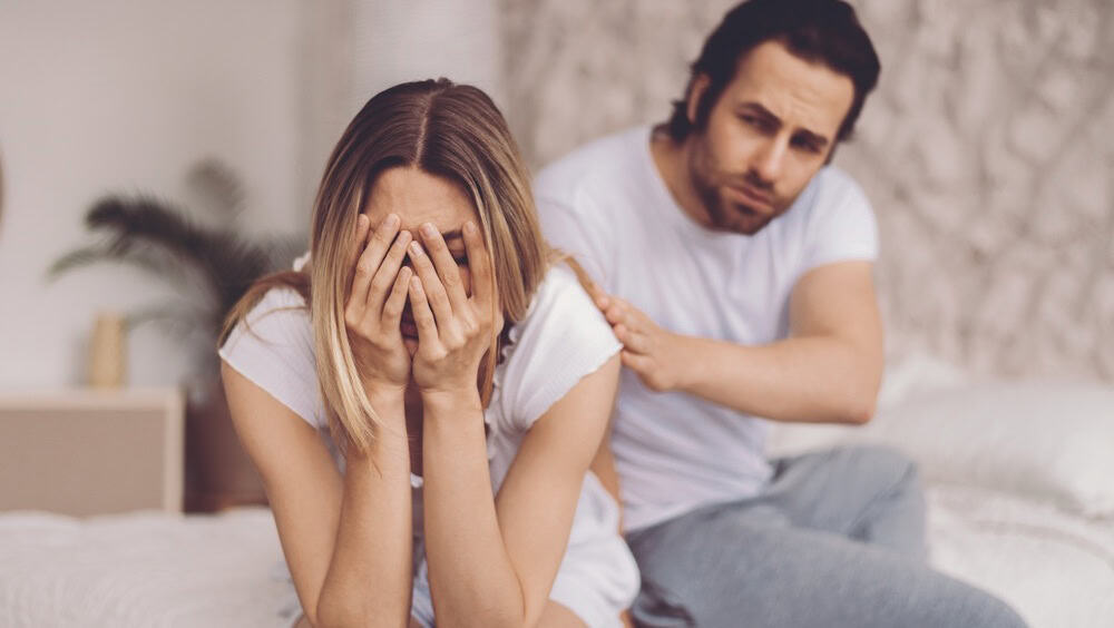 Couple Sitting on Bed With Woman Covering Her Face and Man Showing Concern in a Quiet Room