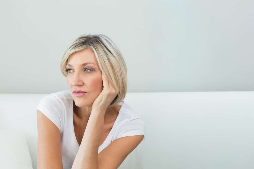 mature woman wearing white top sitting on white couch at home, resting elbow on cushion gazing. Minimalist, neutral, serene, contemplative, thoughtful, settee, adult