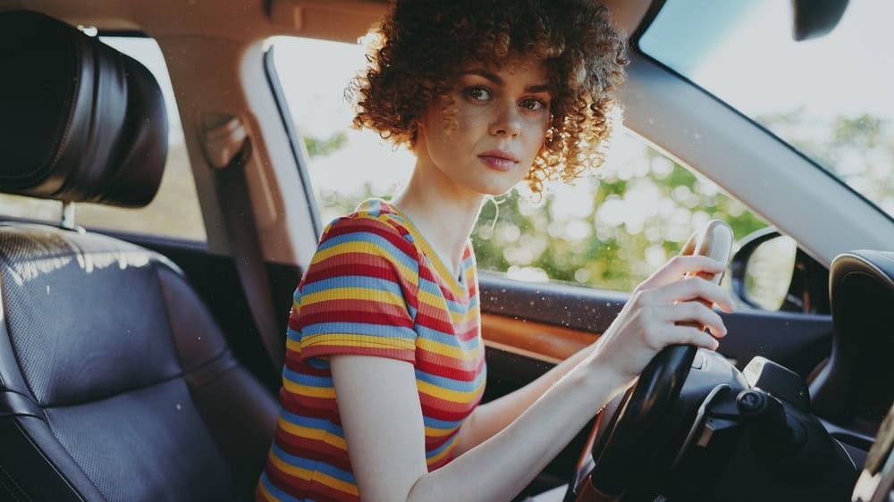 A stylish woman driver sits in a retro car with a striped top, looking at the camera. Warm film colors evoke casual lifestyle and vintage automotive aesthetics.