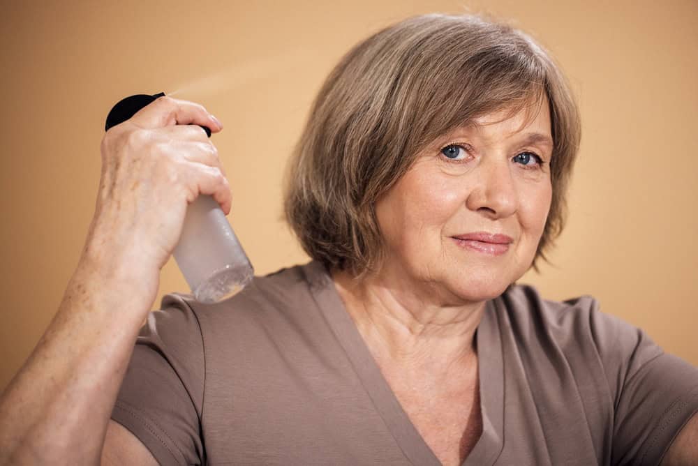 Mature woman with gray hair uses a spray bottle to mist her face, dressed in a beige shirt, with a soft neutral background enhancing the scene