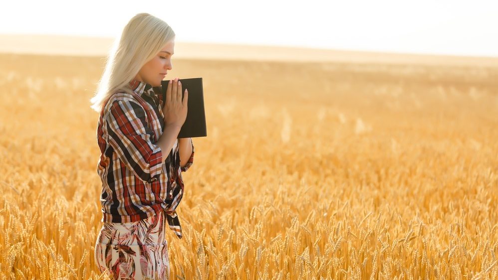 Christian woman praying on holy bible and wooden cross in barley field on summer. Woman pray for god blessing to wishing have a better life and believe in goodness.