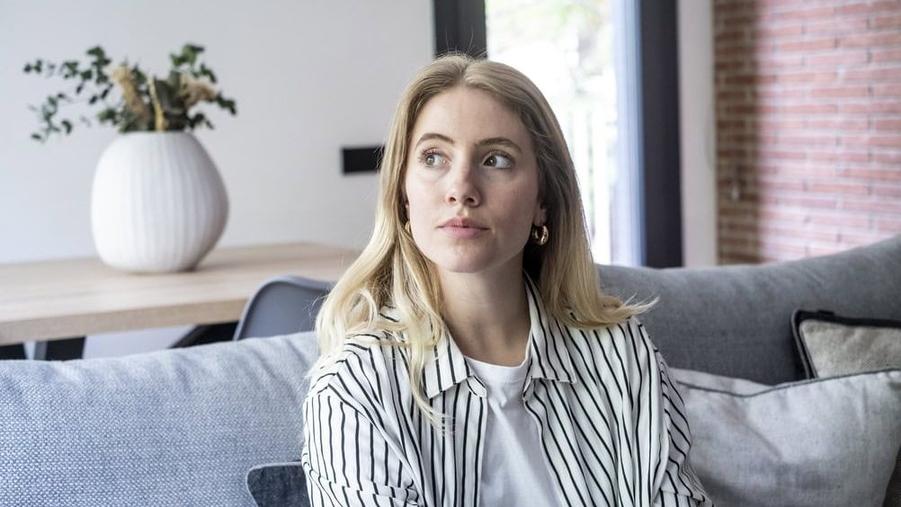 Young blonde woman sitting comfortably on a gray sofa in her modern living room, looking away thoughtfully while contemplating and reflecting