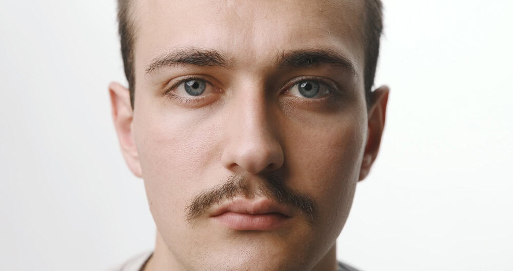 Extreme close-up frontal portrait of a serious young Caucasian man with blue eyes and a neat mustache, looking directly at the camera on a seamless white studio background.