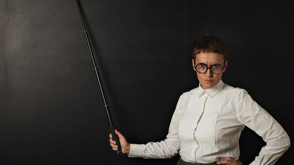 Angry attractive woman teacher looks at camera and shows on black chalk board behind her with folding pointer