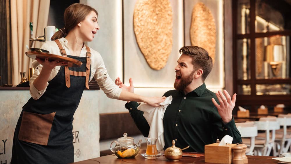 Image of angry screaming bearded young man sitting in cafe. Looking at waiter.