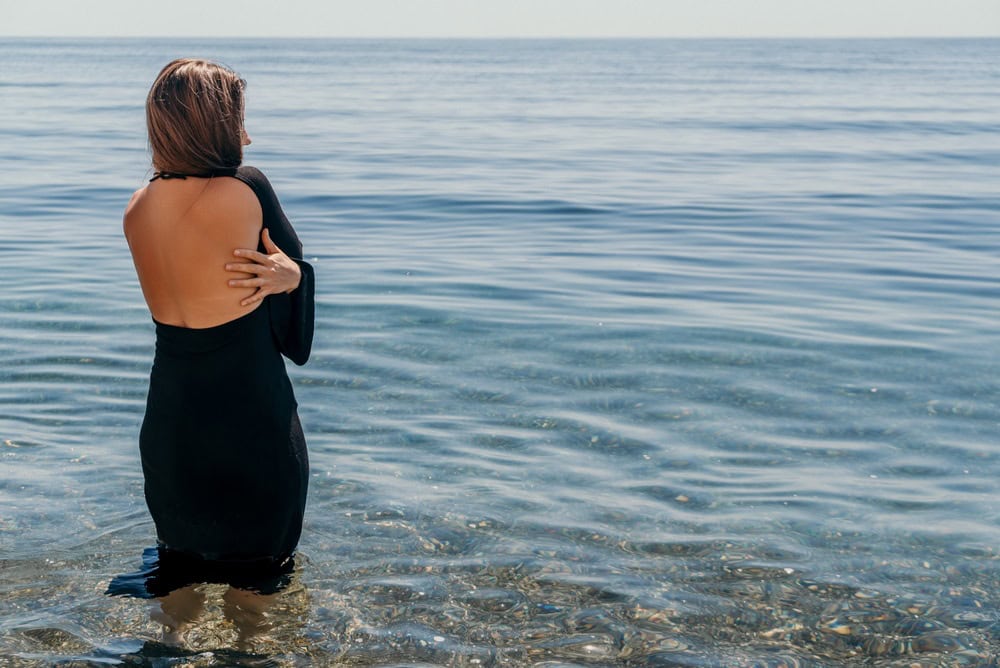 Woman beach ocean brunette in black backless dress standing in clear water while looking at blue sea horizon during summer holiday