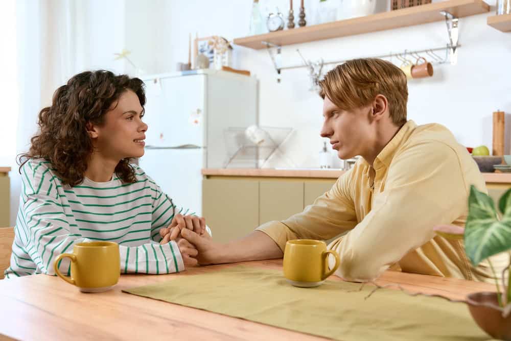 A young couple sits closely at home, sharing an intimate conversation while holding hands
