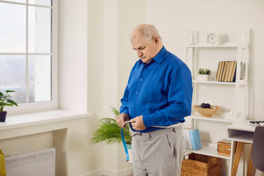 Senior man measuring waist with tape at home. Elderly overweight man checking out his body measuring big stomach. Lose weight, slimming, dieting, health care concept