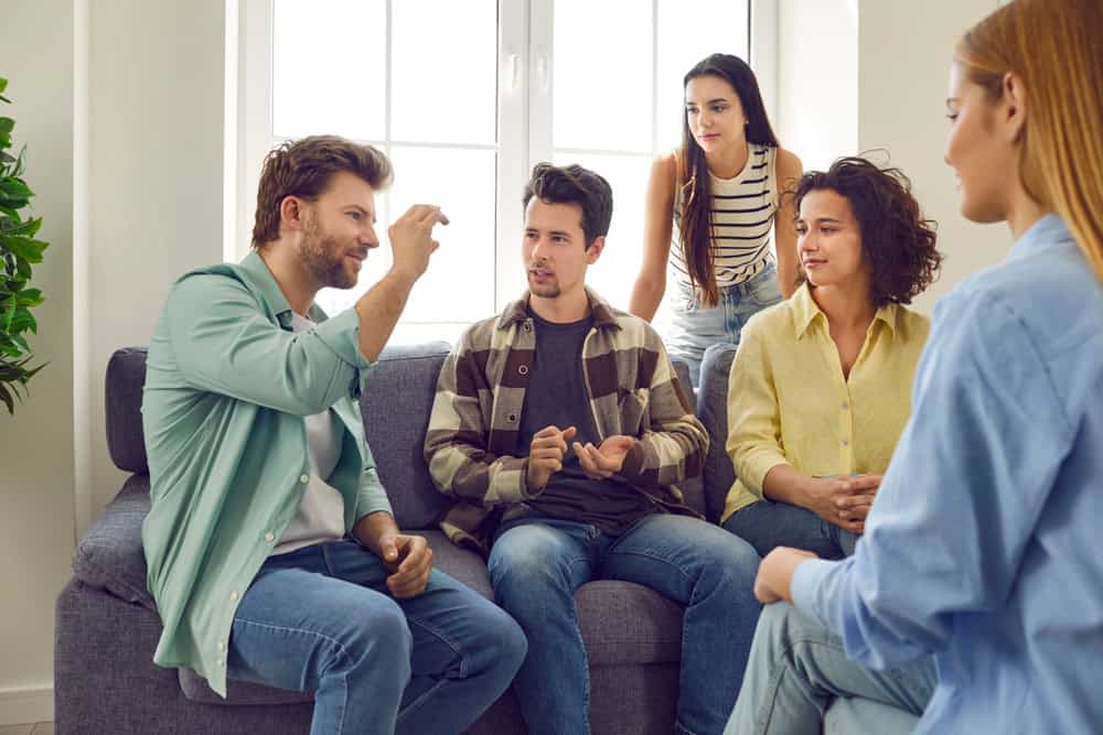 Group of friends gather at home and spend time together. Several people sitting on the couch and talking. Bunch of students playing charades. Young man tells a story or explains a word to his pals