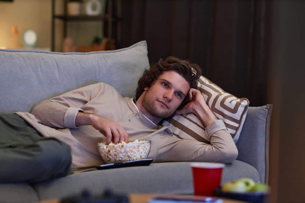 Portrait of young man watching Tv at home and eating popcorn while lying on couch, copy space