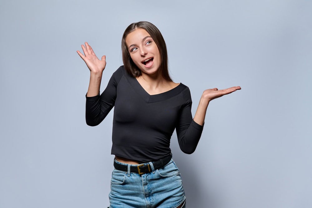 Playful young woman shrugging shoulders and looking away on gray background