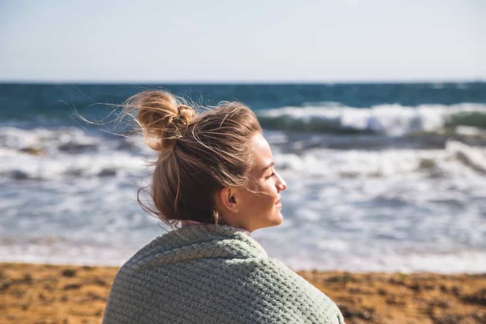 A young beautiful Caucasian girl with a bun of hair on her head in a white boho jacket is sitting on the sand by the sea. Enjoying travel and nature concept.