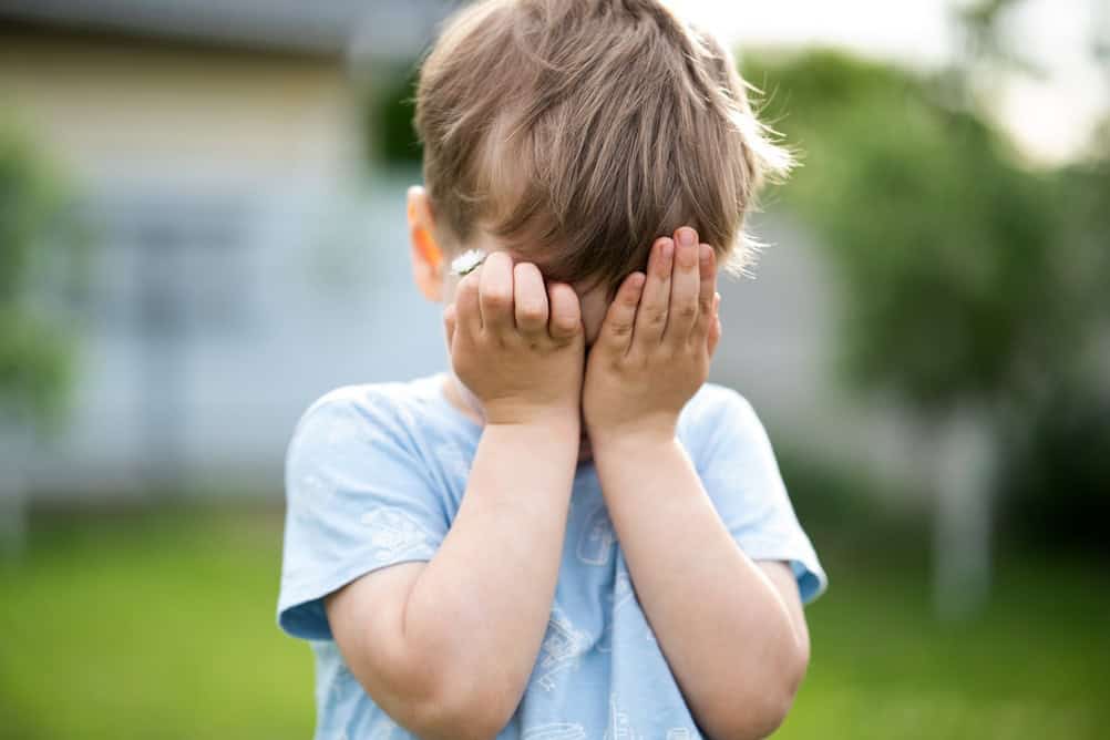 Shy little boy covering his face with hands while holding a daisy flower in a sunny garden. Childhood emotions, innocence, playfulness, outdoor lifestyle and tender moments.