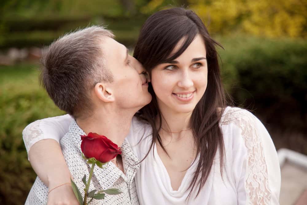 Portrait of couple with red rose on date in park, young man kissing his girlfriend in her ear while