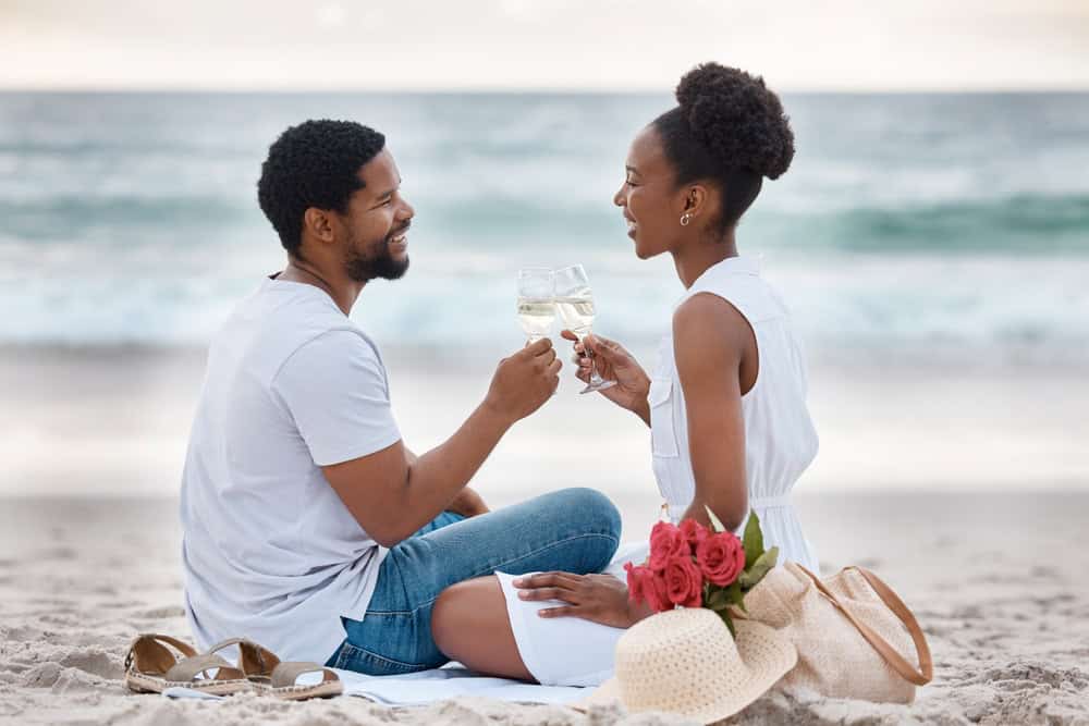 Black couple, toast and relax on sand by beach for romantic date on vacation, anniversary getaway an