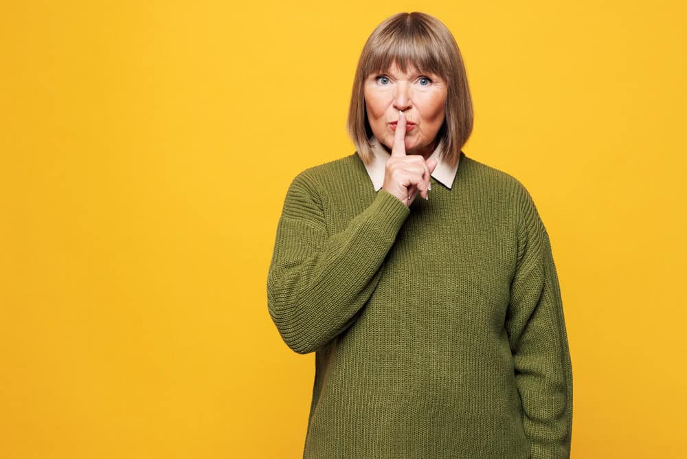 Elderly secret blonde woman she wearing green sweater casual clothes say hush be quiet with finger on lips shhh gesture isolated on plain yellow orange background studio portrait. Lifestyle concept