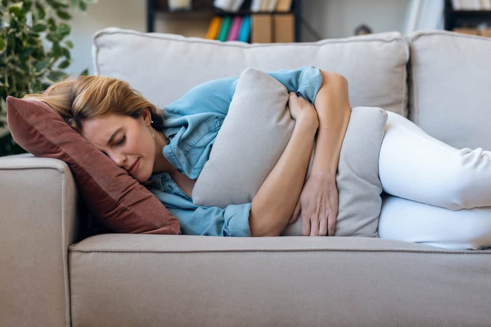 Shot of young tired woman relaxing on couch with pillow for stomachache in living room at home.