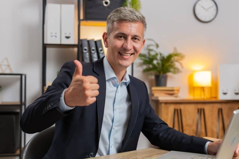 Like. Happy Caucasian businessman looking approvingly at camera showing thumbs up sign positive something good great news positive feedback. Young handsome guy sitting at home office room at table.