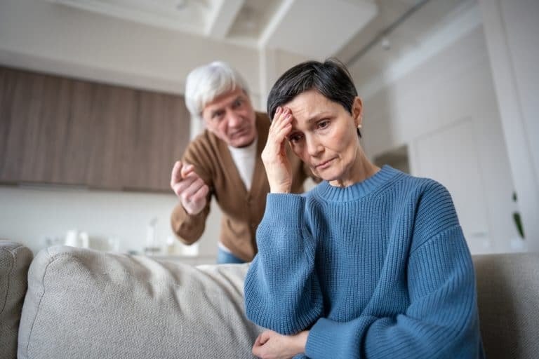 Distressed mature wife hiding from yelling husband, enduring aggression from neurotic man in mental instability. Caregiver stress, manipulative control, suppressed emotions, senior relationship crisis