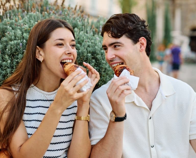 Happy couple eating a pastry croissant, smiling and looking away on a city street
