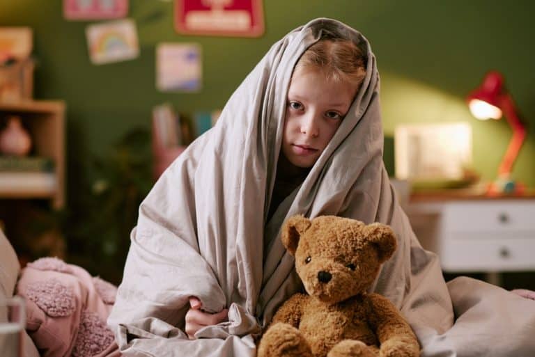 Portrait of Caucasian child girl sitting on bed wrapped in blanket holding toy bear, looking at camera with serious expression bedroom background with toys and decorations visible
