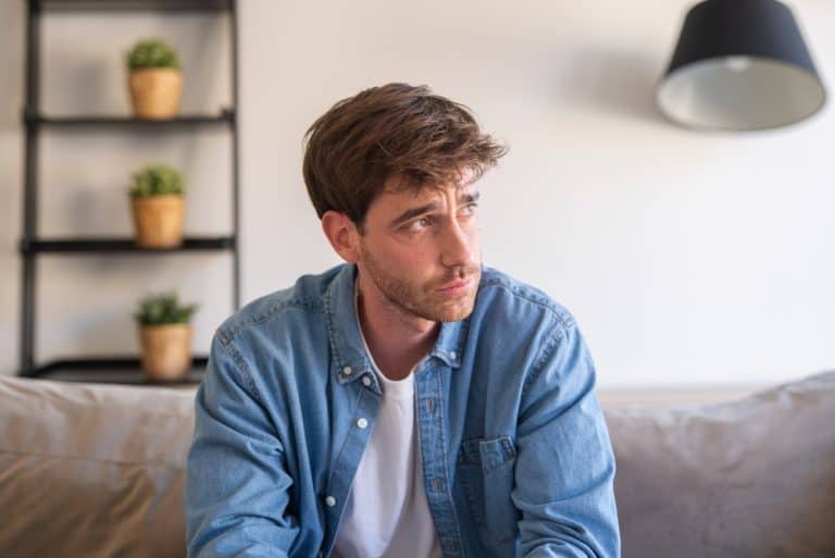 An upset millennial man sits alone on a sofa, thinking about personal problems and feeling anxious and disconnected in a silent home environment